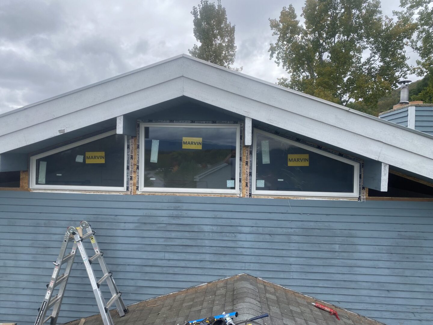 Three triangular windows installed on a blue house under a cloudy sky.