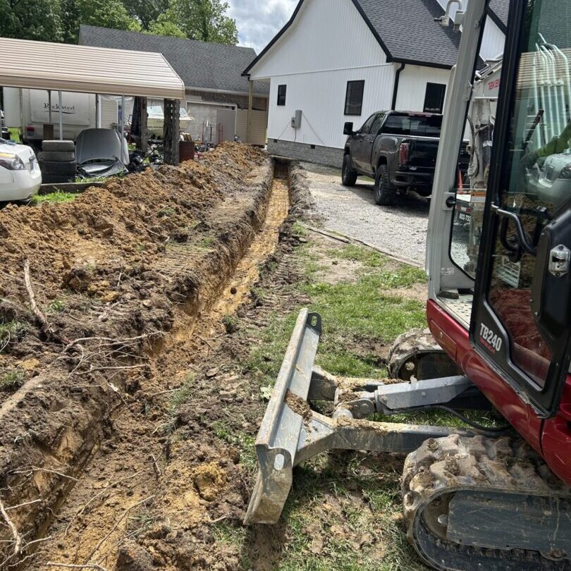 Excavator digging a trench near a house with parked cars.